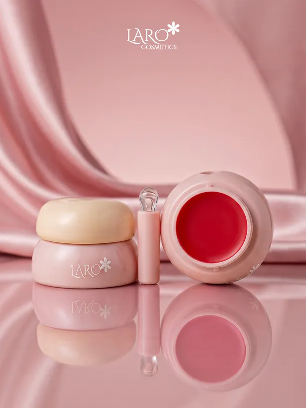 Product photograph: an open red lip and cheek tint pot with its applicator, reflected on a glossy surface.
